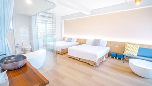 Egy modern szállodai szoba, melyben üvegfalas zuhanyzó, fa mosdó, nagy, fehér ágyneművel ellátott ágy, kék kanapé és kör alakú, fehér dohányzóasztal található.