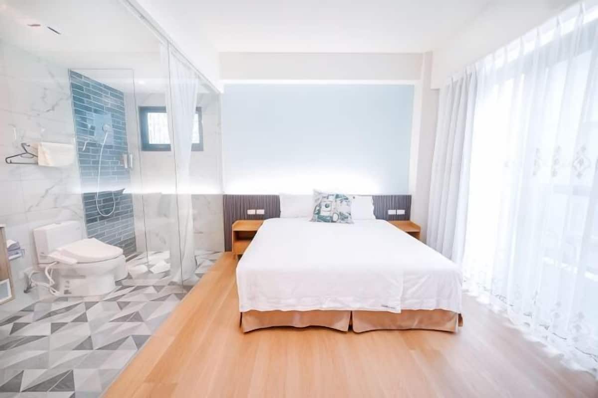 A modern hotel room with a large bed, a glass shower enclosure, and a view of the city.