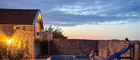 A swimming pool with a slide, a stone wall, and a building with a light.