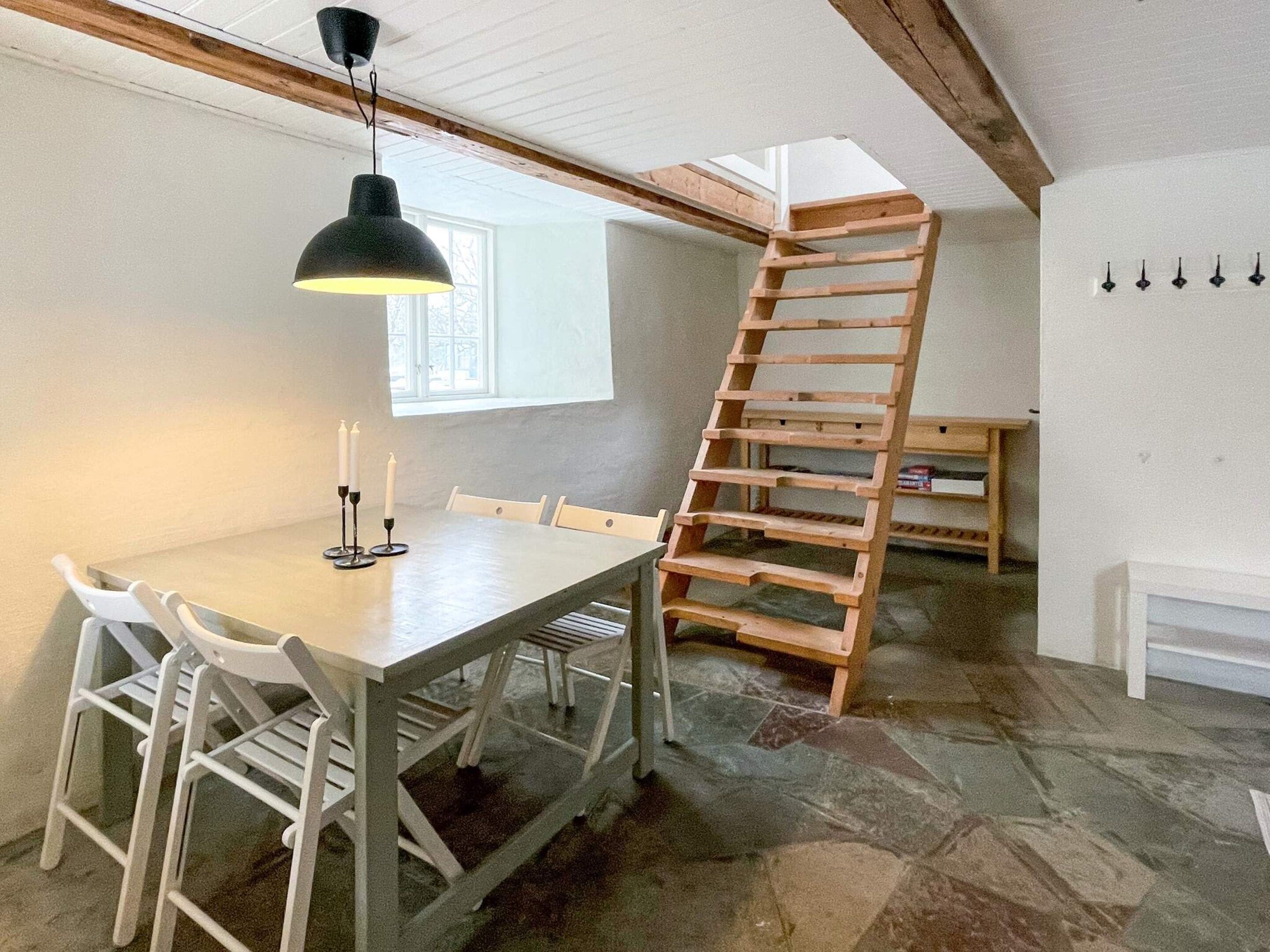 Un comedor con una mesa, sillas, una lámpara colgante, una escalera de madera y una ventana.