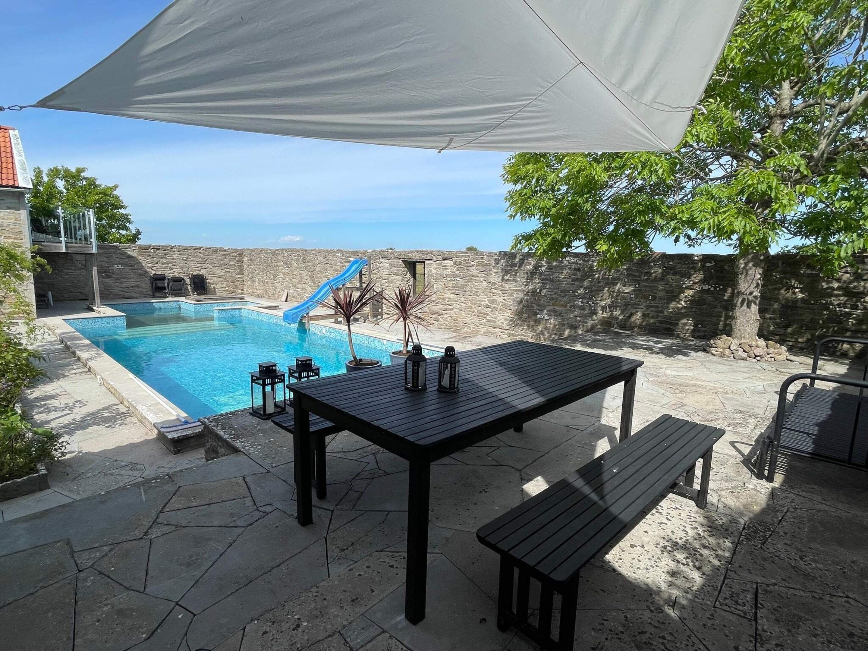 Ein Poolbereich mit Tisch und Bänken, umgeben von Steinmauern und blauem Himmel.