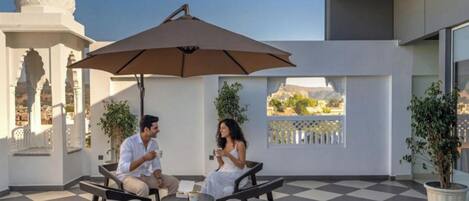 A terrace with a checkered floor, outdoor seating, a large umbrella, and a view of a white building with domes.