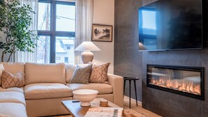 Ein modernes Wohnzimmer mit einem beigefarbenen Sofa, einem Couchtisch mit einem Buch, einem Kamin und einem großen Fenster mit Vorhängen.