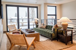 A cozy living room with a green sofa, wicker chairs, a wooden coffee table, and a small side table with a lamp. - Luxe Downtown Penthouse • Panoramic Hot Tub Views (Whitefish)