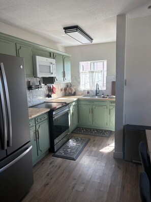 A kitchen with green cabinets, a stainless steel refrigerator, a microwave, and a window with curtains.