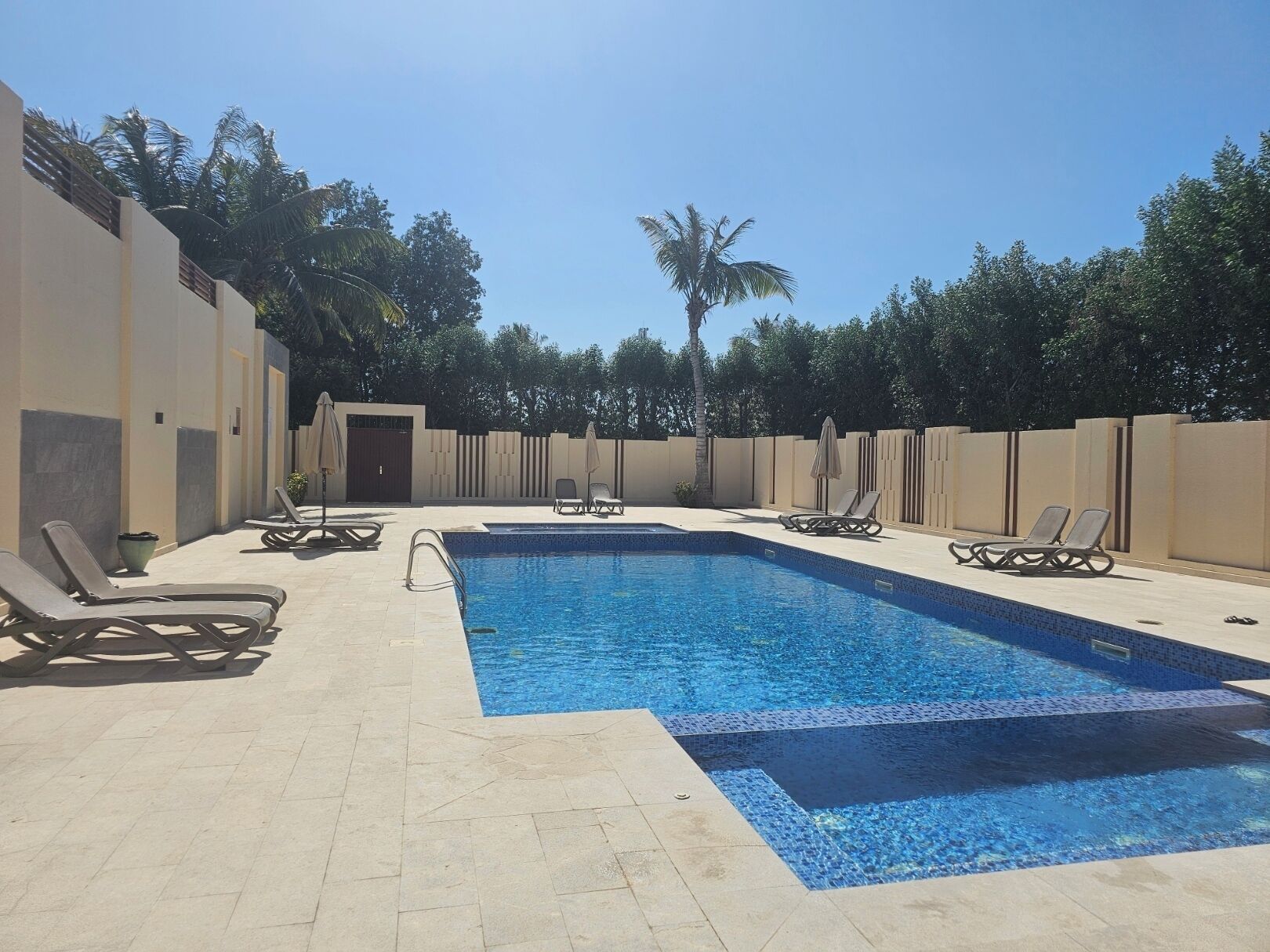 Une piscine rectangulaire bordée de carreaux, entourée de murs de couleur beige et dotée d’un sol carrelé. Des chaises longues et des parasols sont disposés le long du bord de la piscine.