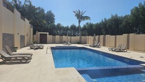 Una piscina rectangular con borde azulejado, rodeada de paredes color beige y suelo de baldosas. Hay sillas de descanso y sombrillas a lo largo de la piscina.