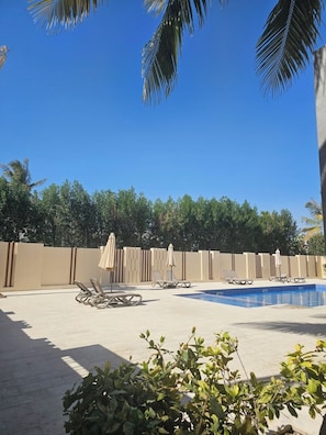 A pool area with lounge chairs and umbrellas, surrounded by a low wall and palm trees. - Hawana Salalah - Private studio with mountain view and swimming pool (Salalah)