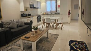 A modern apartment with a living room, dining area, and kitchen. There is a sofa, a coffee table, and a dining table with chairs. The kitchen has a microwave, oven, and sink.