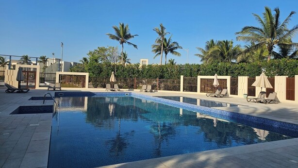 A swimming pool with tiled edges, surrounded by lounge chairs and umbrellas, with palm trees and a clear sky in the background.