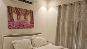 Un dormitorio con cama, mesita de noche, aire acondicionado, un cuadro y una ventana con cortinas.