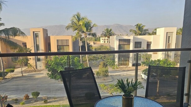 Un balcón con una mesa redonda y cuatro sillas, con vista a una calle con edificios y palmeras.