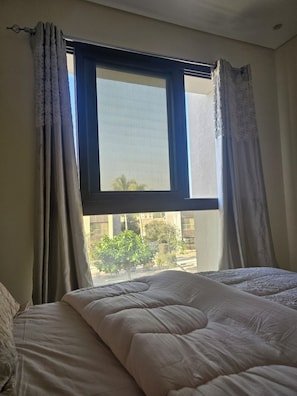 A room with a bed, a window with curtains, and a view of buildings and trees outside. - Hawana Salalah - Private studio with mountain view and swimming pool (Salalah)