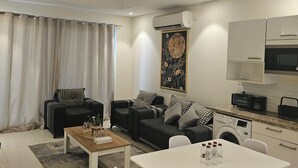 Una sala moderna con un conjunto de sofás, una mesa de centro y una mesa de comedor con sillas. Hay una zona de cocina con microondas y lavadora.