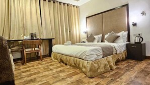 A hotel room with a bed, a desk, a chair, a ceiling fan, and a nightstand with a lamp. - Manuhar (Jaipur)