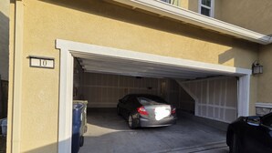 Eine Garage mit einem darin abstellendem Auto, einem Müllbehälter und einem Hinweisschild mit der Hausnummer.