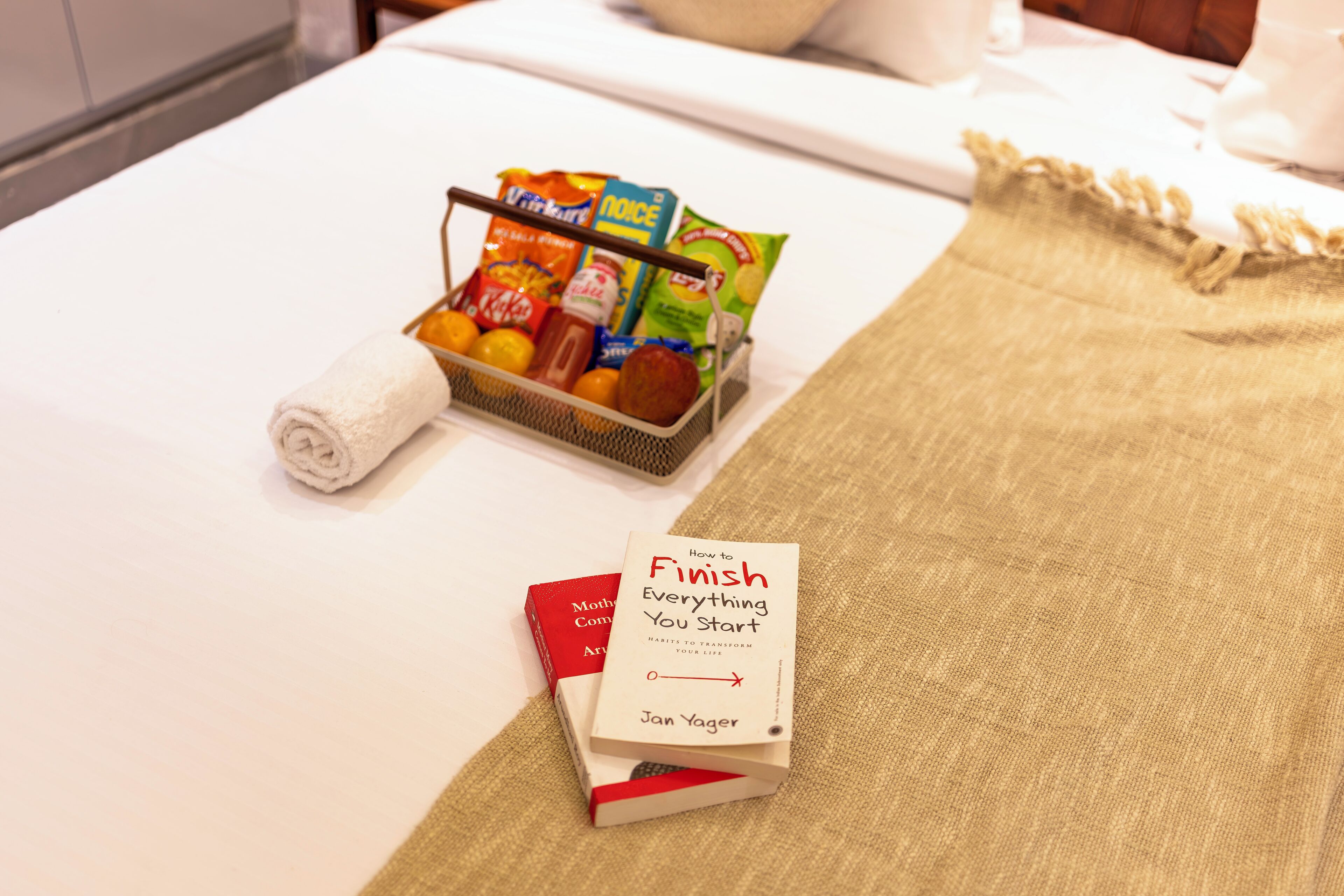 Een net opgemaakt bed met een opgerolde witte handdoek, een mand met snacks en een boek getiteld 