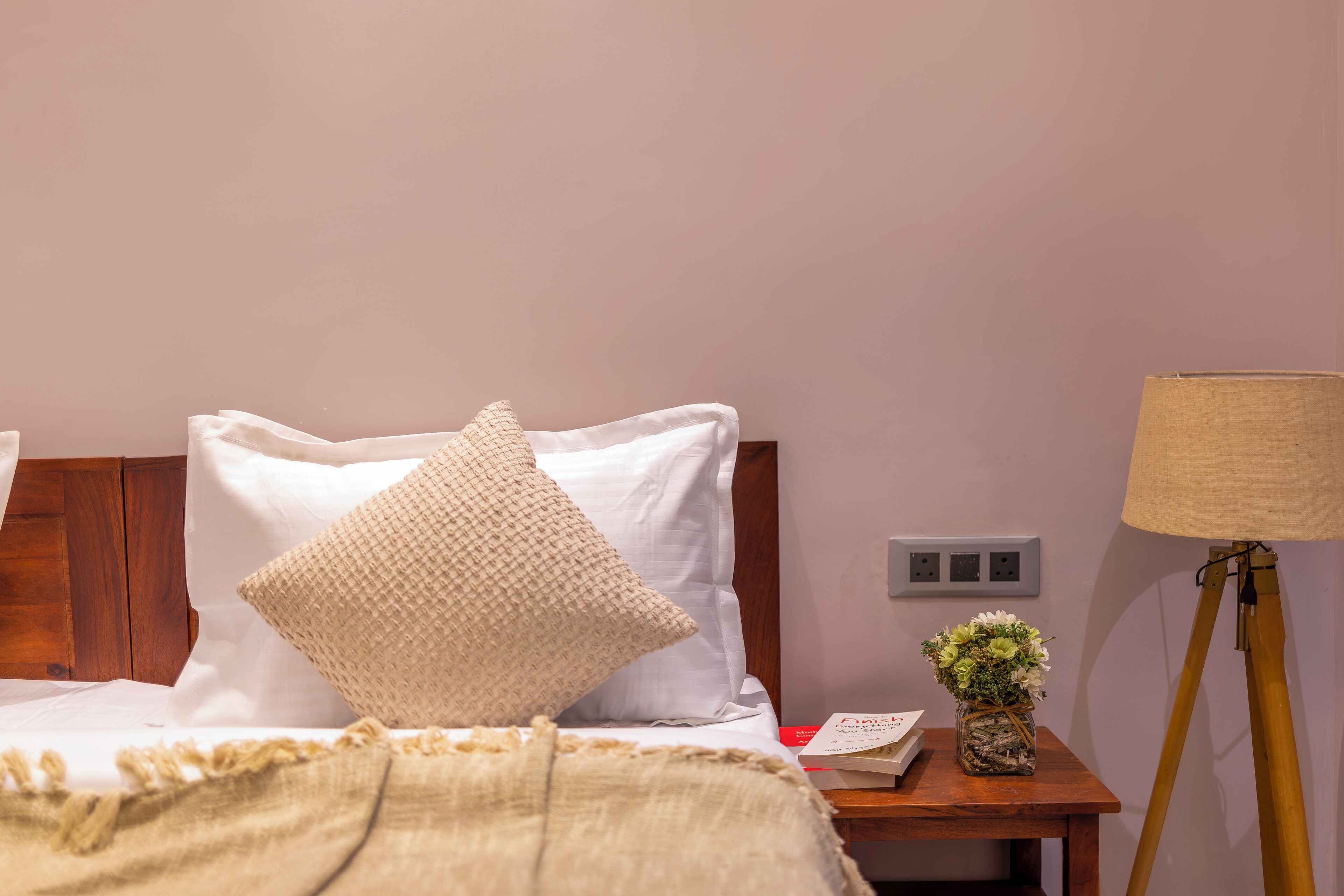 A neatly made bed with white linens and a beige throw blanket, a bedside table with a lamp and a small vase of flowers, and a wooden nightstand.