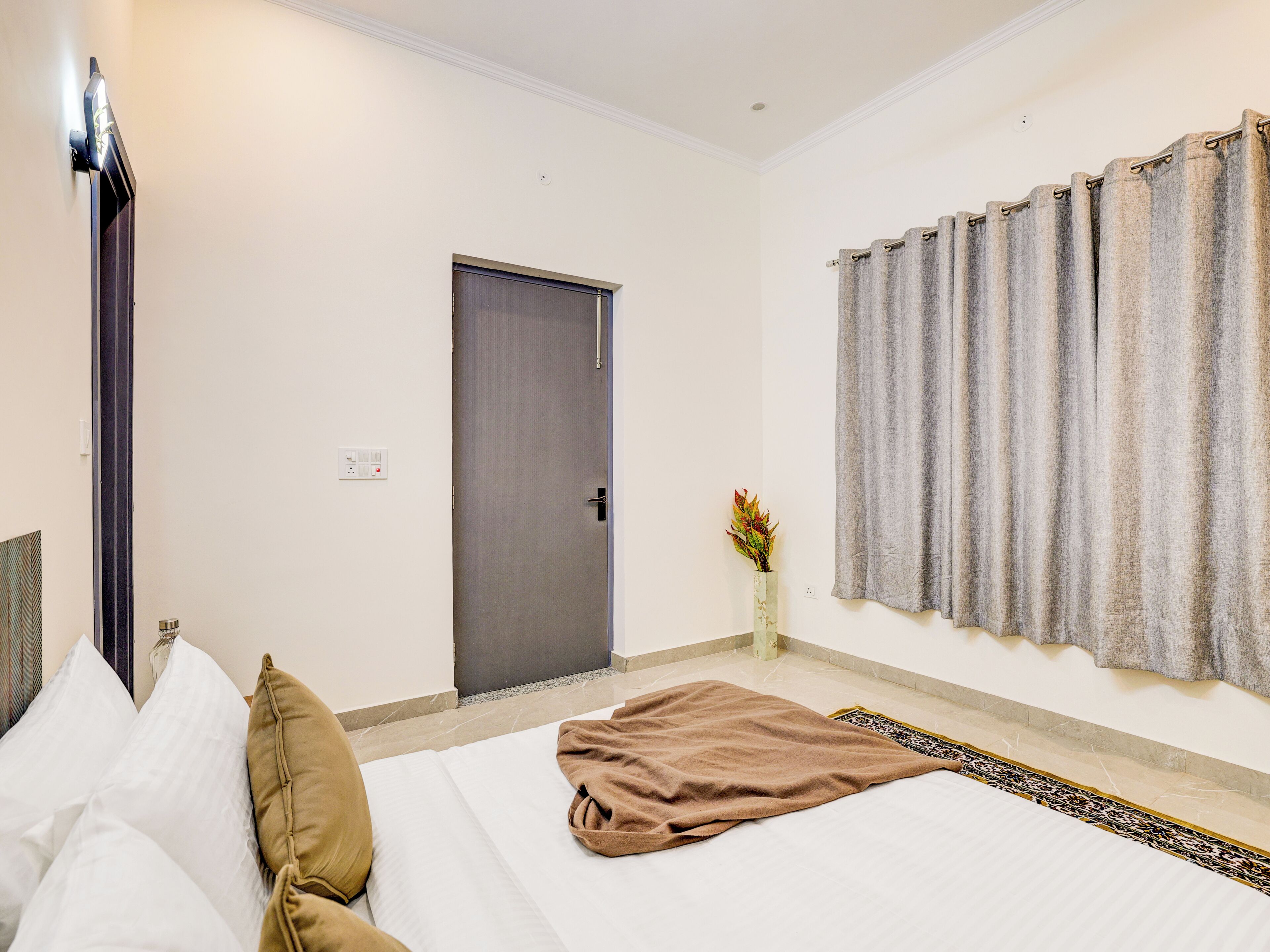 A hotel room with a bed, pillows, a blanket, a door, a curtain, and a vase with flowers.