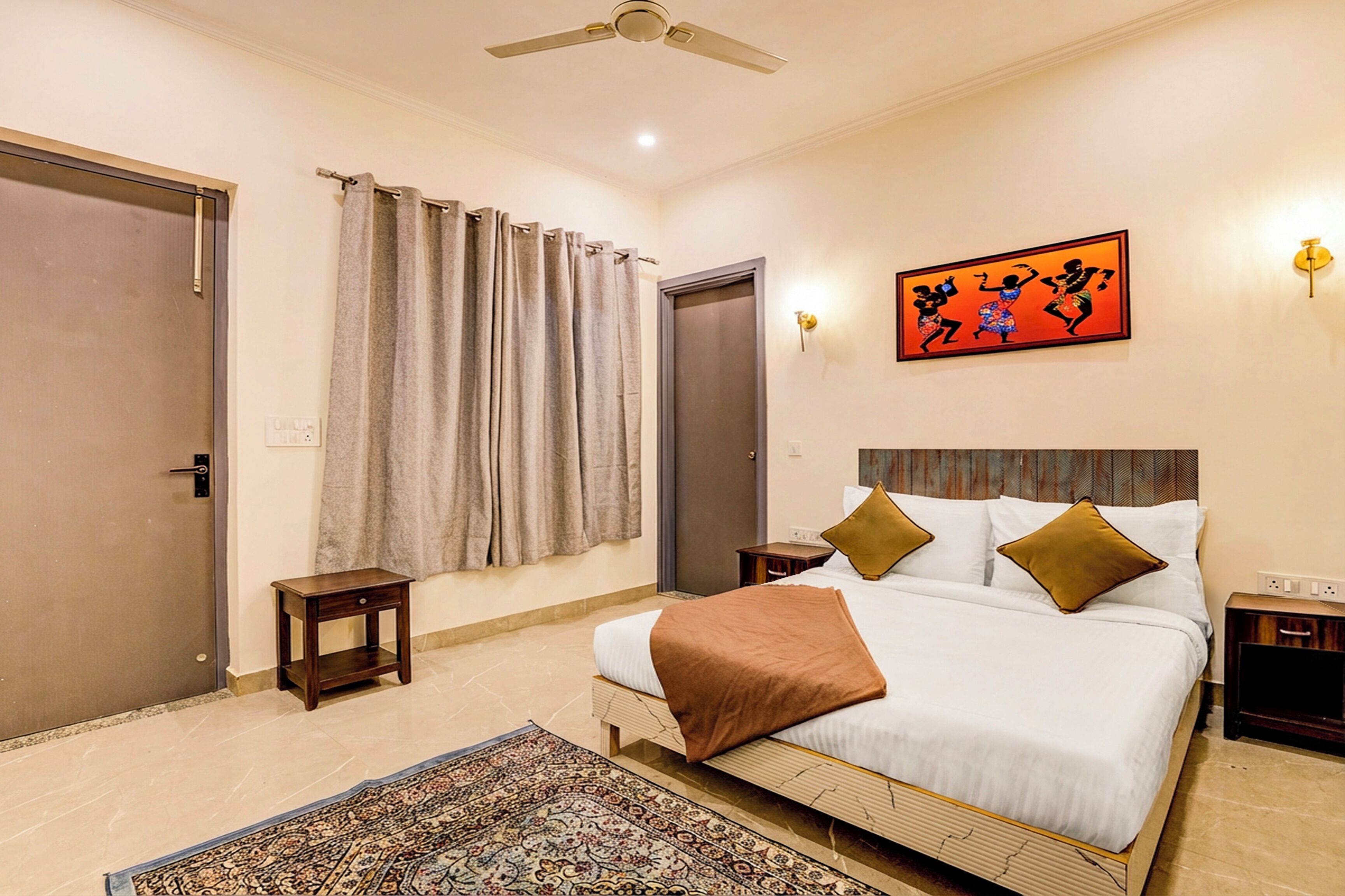 A hotel room with a bed, curtains, a small table, a nightstand, a wall-mounted light, and a ceiling fan.