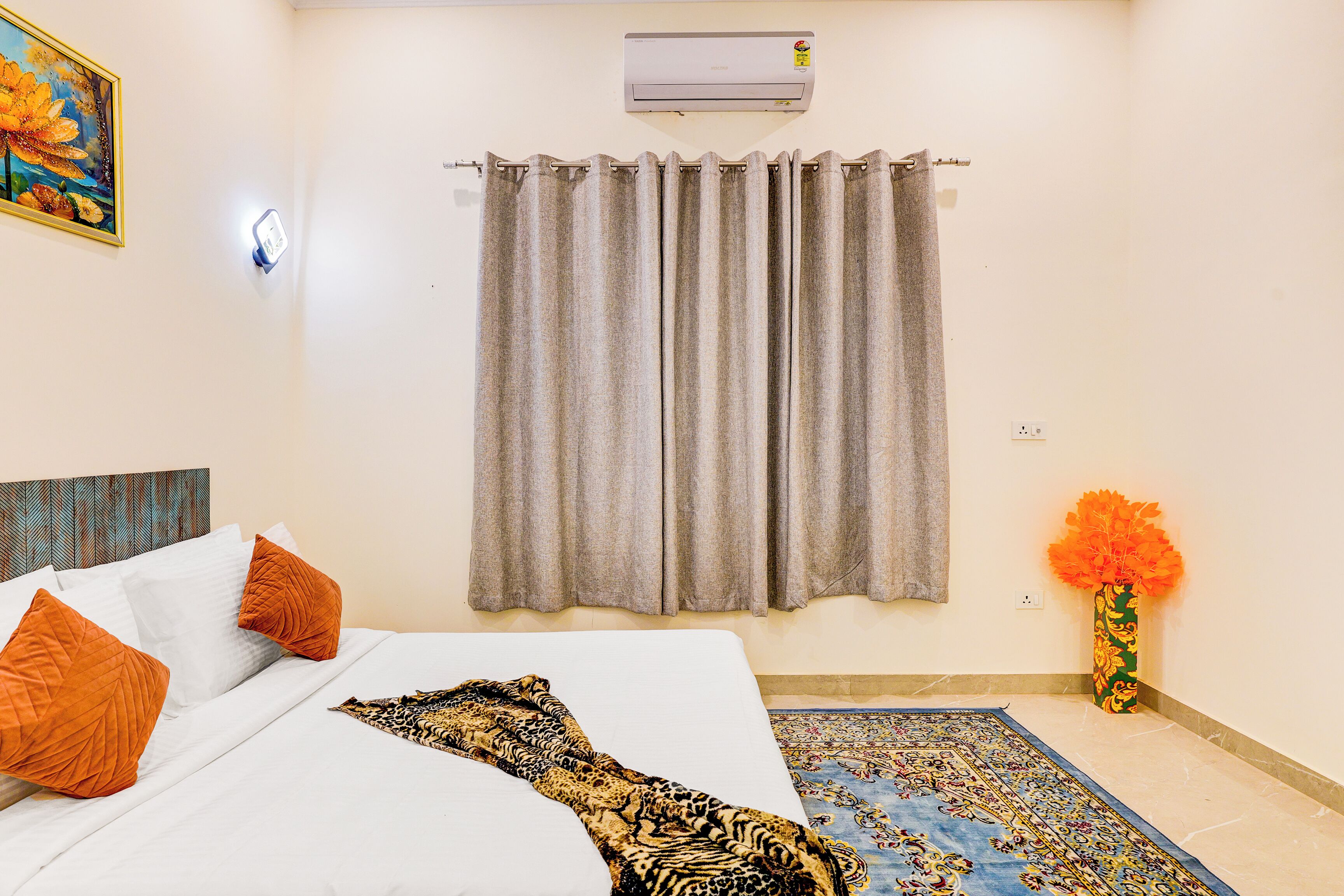 A hotel room with a bed, curtains, a wall-mounted air conditioner, a painting, and a vase with flowers.