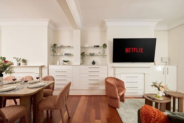A modern living room with a flat-screen TV, a dining area with a wooden table and chairs, and a kitchen area with white cabinetry and shelves.