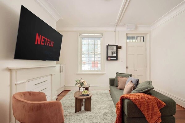 A living room with a flat-screen TV, a sofa, a chair, a small table with a vase, and a window. - 4BR Surry Hills Home | Pet Friendly + Parking (Surry Hills)