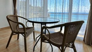 A room with a round glass table and two wicker chairs, large windows with sheer curtains, and a view of the sea. - Casa Mila Apartments & Bungalows (Utjeha)