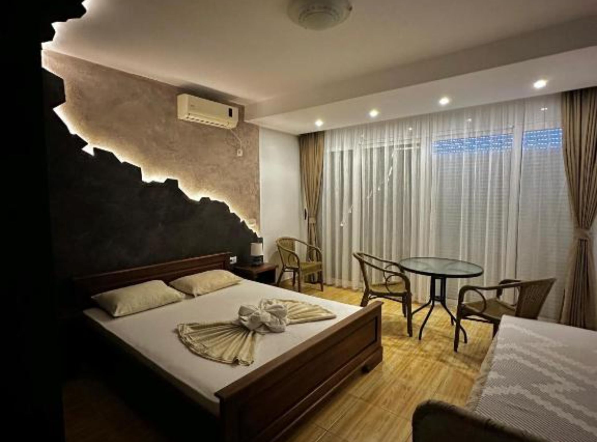 A hotel room with a bed, a small table, and a chair. There is a window with curtains and an air conditioning unit on the wall.