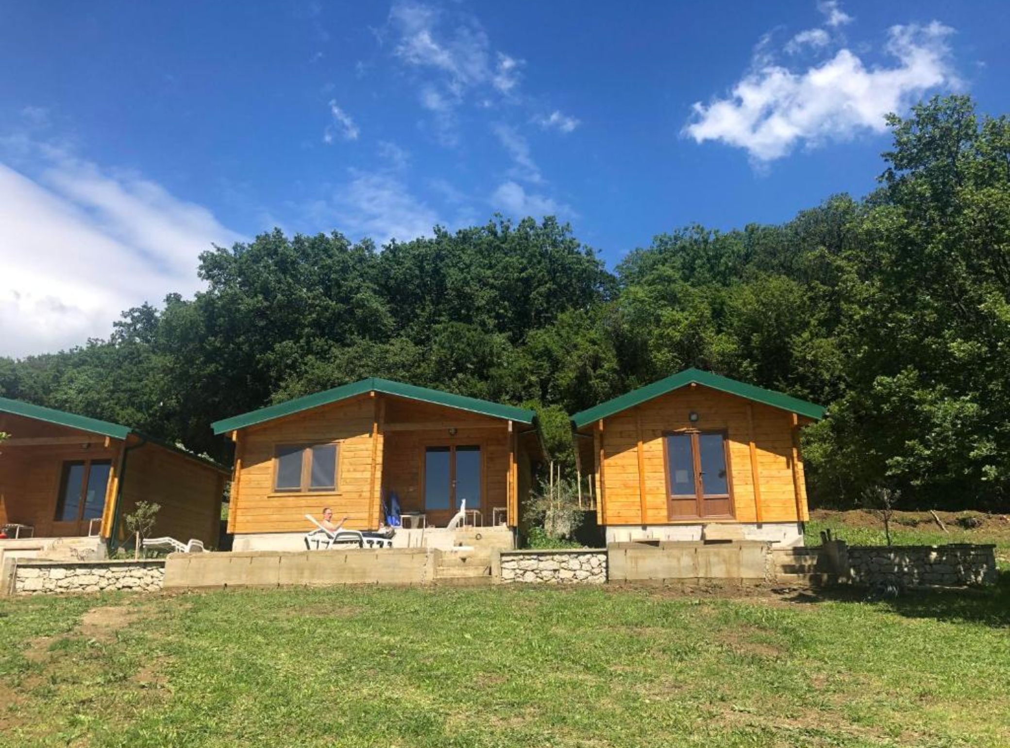 Three wooden cabins with green roofs, set in a grassy area with trees in the background.