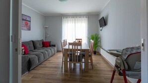 A modern living room with a grey sofa, wooden dining table, and chairs. There is a television mounted on the wall and a window with curtains. - Apartments Slavek (Jastrebarsko)