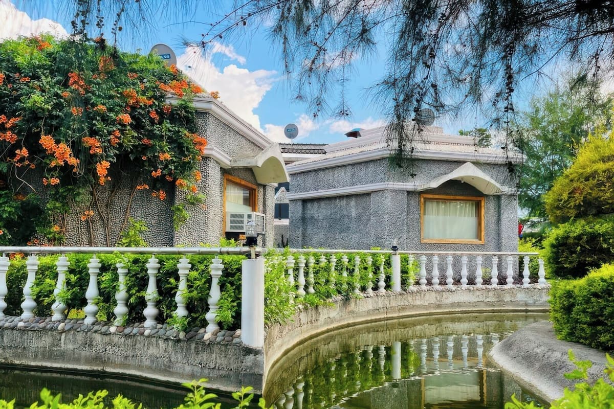 A stone building with a small bridge over a water feature, surrounded by lush greenery and flowering plants.