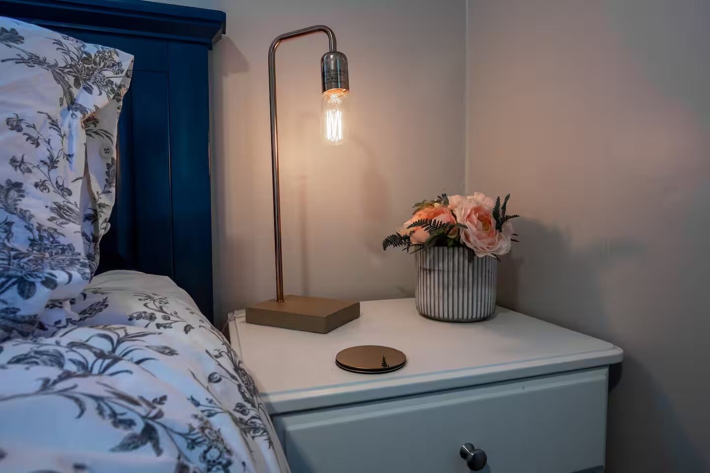 Een netjes opgemaakt bed met bloemenbeddengoed, een bedlampje, een kleine vaas met bloemen en een witte nachtkastje met een lade.