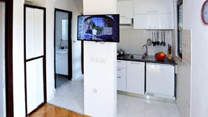 A modern kitchen with white cabinets, a built-in oven, and a flat-screen TV mounted on a white pillar. - Apartments Nena (Kukljica)
