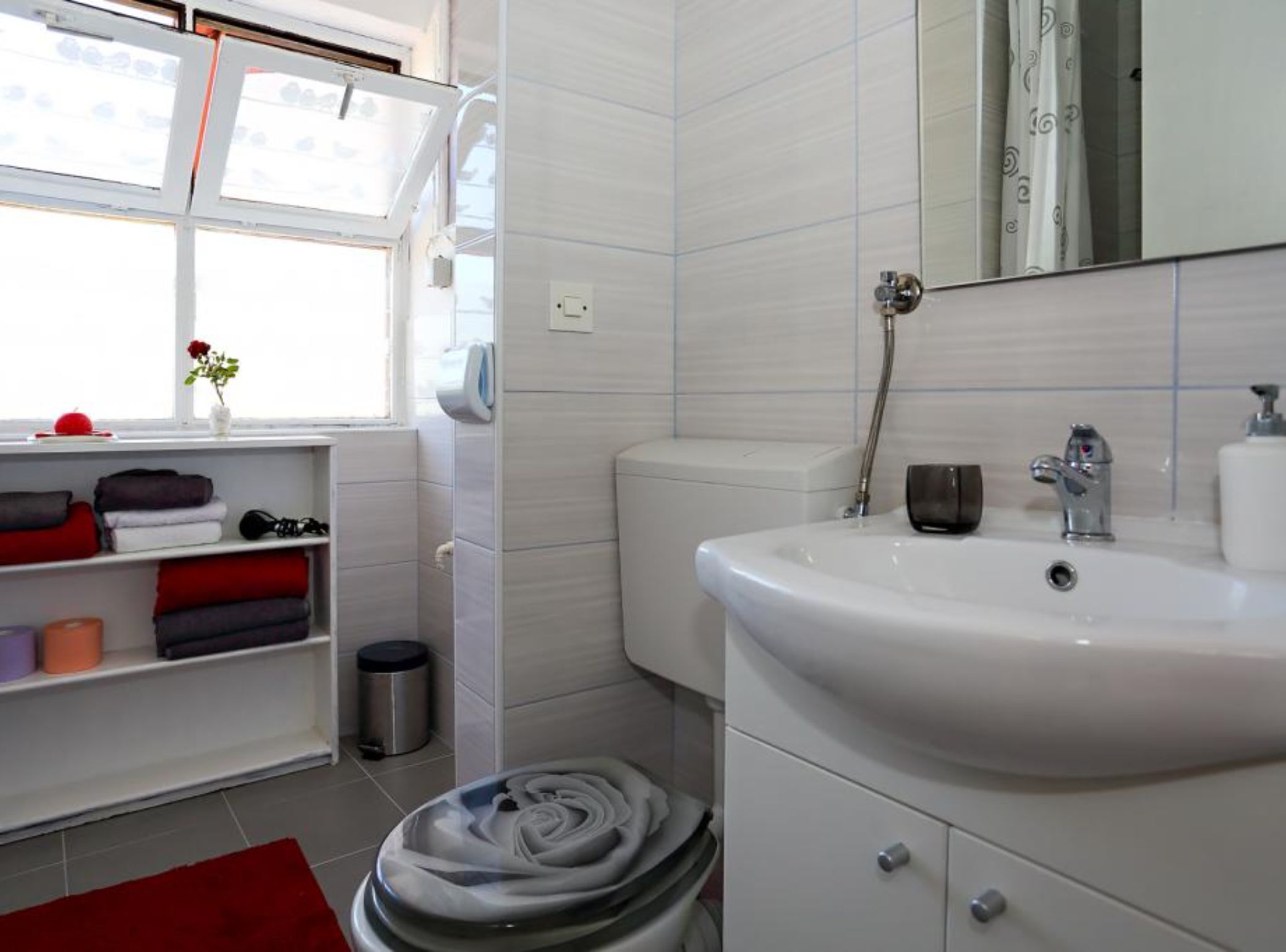 Et moderne badeværelse med hvid vask, toilet og brusebad. Der er et vindue med udsigt, en hylde med håndklæder og en skraldespand.