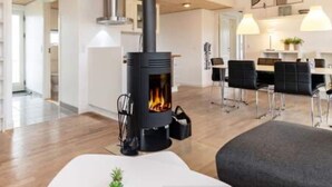 A modern living room with a wood-burning stove, a dining area with chairs and a table, and a kitchen in the background. - 3 bedroom amazing home in Brovst (Brovst)