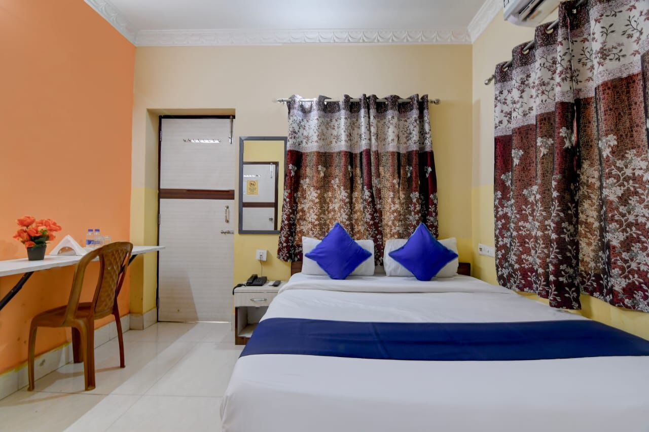 A hotel room with a bed, a desk, and a chair. There is a door and a nightstand with a lamp.