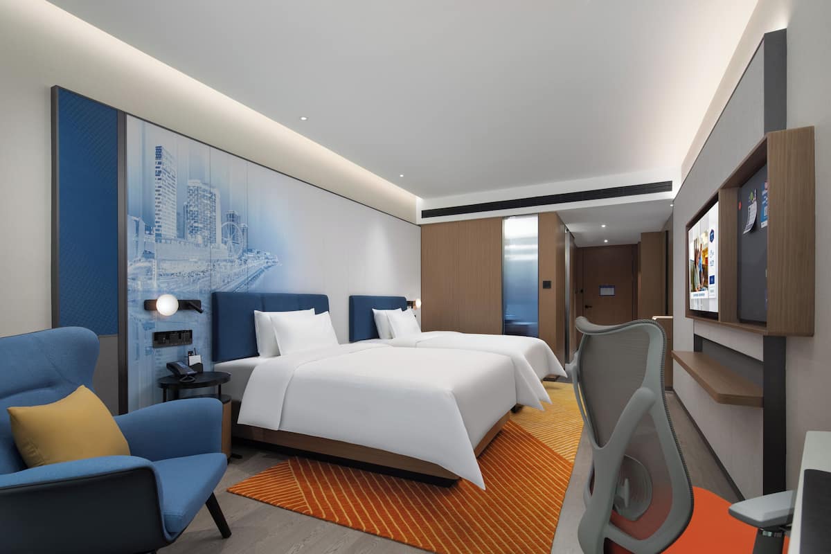 A modern hotel room with a large bed, a desk, a chair, a sofa, and a cityscape mural on the wall.