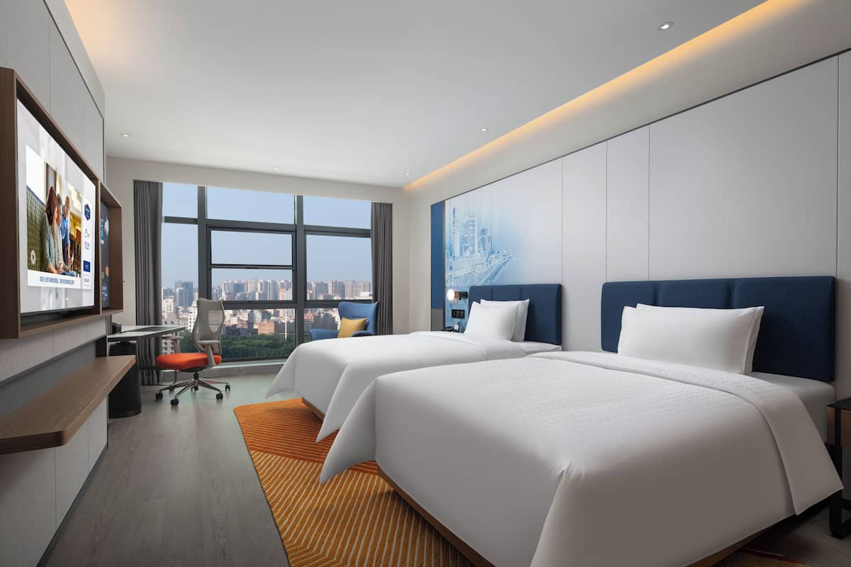 A hotel room with two beds, a desk, a chair, and a large window offering a city view.