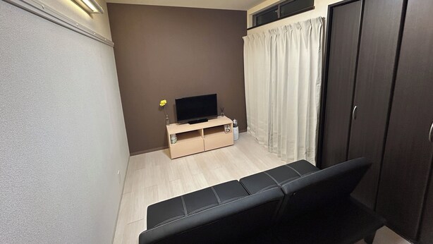 Una habitación pequeña y funcional con un banco, un televisor de pantalla plana sobre un soporte de madera, un armario y una ventana con cortinas.