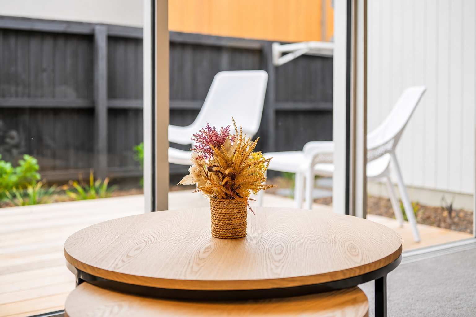 A round wooden table with a woven basket containing dried flowers, set against a backdrop of a wooden fence and white chairs.