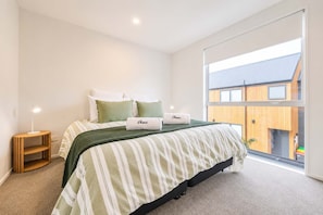 A bedroom with a bed, a bedside table, a lamp, and a window with a view of a building. - Two Storey 2BR | Private Patio | Near CBD (Christchurch)