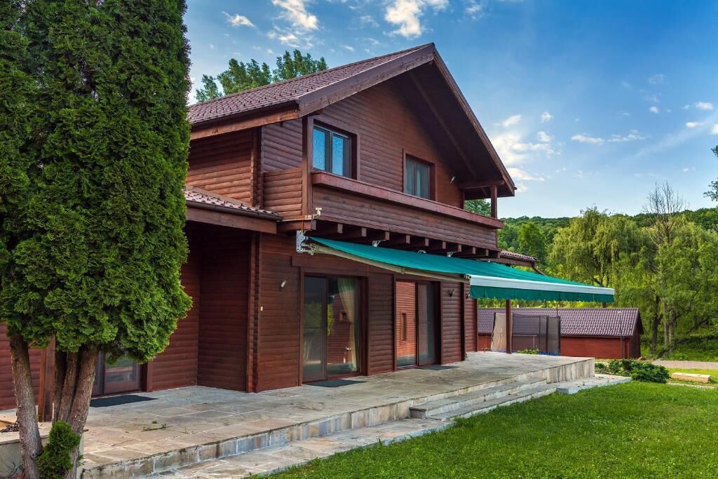 A two-story wooden cabin with a green awning, a stone-paved entrance, and a well-maintained lawn.
