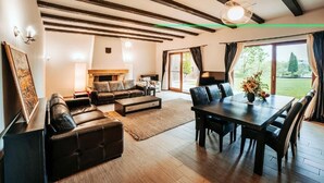 Ein modernes Wohnzimmer mit einer Couch, einem Couchtisch und einem Esstisch. Ein Kamin, ein großes Fenster und eine Decke mit sichtbaren Balken sind ebenfalls vorhanden.