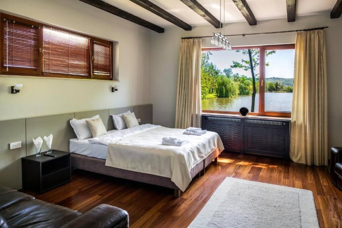 A bedroom with a large bed, wooden flooring, a view of a lake, and a window with curtains.