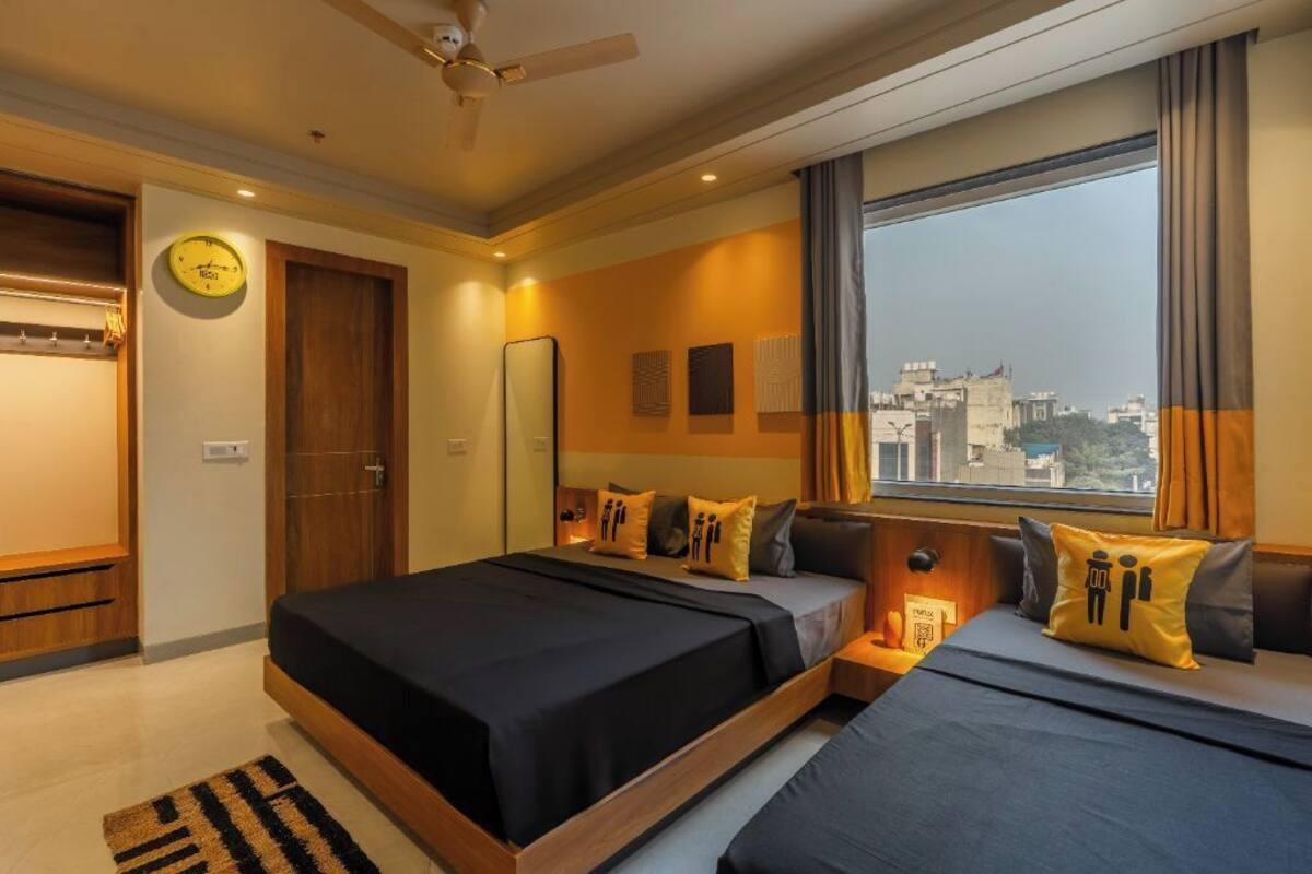 Une chambre d’hôtel avec deux lits, un ventilateur de plafond, une grande fenêtre avec des rideaux, une horloge au mur et une vue sur la ville.