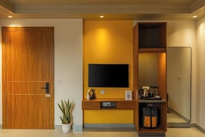 A hotel room with a wooden door, a television mounted on a yellow wall, a small shelf with a coffee maker, and a mirror on the right wall. - The Hosteller Delhi, International Airport (New Delhi)