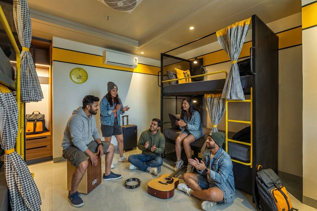 A group of people in a room with bunk beds, a clock on the wall, and a guitar on the floor.