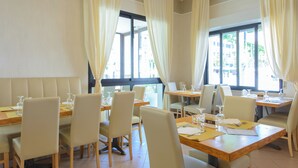 Un comedor con mesas y sillas, ventanas amplias con cortinas, y vistas a edificios.