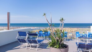 A terrace with blue chairs and a potted plant, overlooking a beach with umbrellas and people. - Hotel Nuova Medusa (Rimini)
